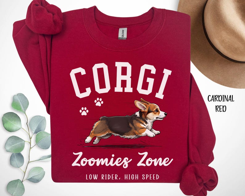 May include: Cardinal red sweatshirt with the word "CORGI" in white block letters, a graphic of a running corgi, and the text "Zoomies Zone: Low Rider, High Speed". The sweatshirt has a crew neck and long sleeves.