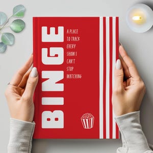 May include: A red journal with the word "BINGE" in large white letters. The journal also features the text "A place to track every show I can't stop watching" and a popcorn graphic. White vertical stripes are on the right side.