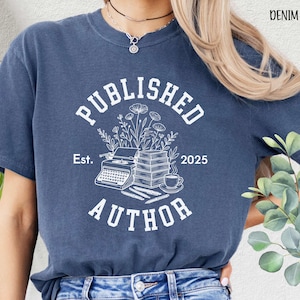 May include: Denim blue t-shirt with a white graphic that reads "PUBLISHED AUTHOR" with a typewriter, books, flowers, and a coffee cup. The text "Est. 2025" is also included in the design. The shirt is a casual style.