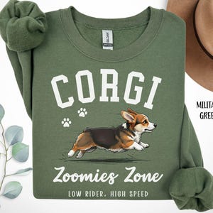 May include: Military green sweatshirt with the word "CORGI" in white block letters above a running corgi illustration. Below the illustration, the text reads "Zoomies Zone, LOW RIDER, HIGH SPEED". The sweatshirt has a crew neck and long sleeves.