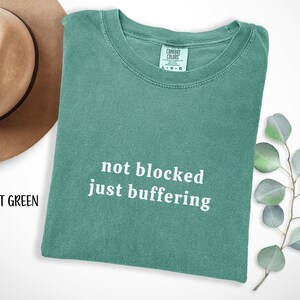 May include: A light green t-shirt with the text "not blocked just buffering" in white. The shirt is folded and lies on a white surface. A tan hat is partially visible in the upper left corner.