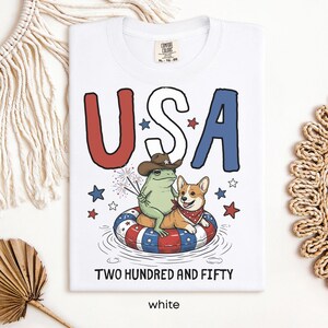 Puede incluir: Camiseta blanca con un dise&ntilde;o patri&oacute;tico que presenta la palabra "USA" en rojo y azul, una rana con sombrero de vaquero y un corgi con pa&ntilde;uelo. La camiseta tambi&eacute;n tiene el texto "TWO HUNDRED AND FIFTY" y "white".