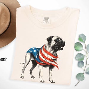 May include: Ivory t-shirt with a graphic of a Bullmastiff dog wearing a red, white, and blue American flag bandana. The text "COMFORT COLORS" is printed on the tag.