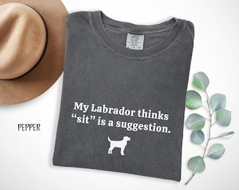 Puede incluir: Camiseta gris oscuro con el texto "My Labrador thinks "sit" is a suggestion." y una silueta blanca de perro. Un sombrero marrón está en la esquina superior izquierda. La palabra "PEPPER" está en la esquina inferior izquierda.