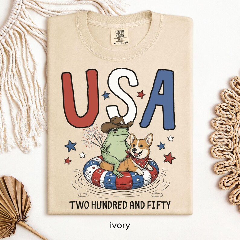 Puede incluir: Camiseta de color marfil con la palabra "USA" en rojo, blanco y azul. Una rana con sombrero de vaquero y un corgi con pa&ntilde;uelo flotan en una c&aacute;mara de aire. El texto "TWO HUNDRED AND FIFTY" est&aacute; debajo.