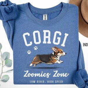 May include: Indigo blue sweatshirt with the word "CORGI" in white block letters above a running corgi illustration. The text "Zoomies Zone" and "LOW RIDER, HIGH SPEED" are printed below the dog. Paw prints are also included.