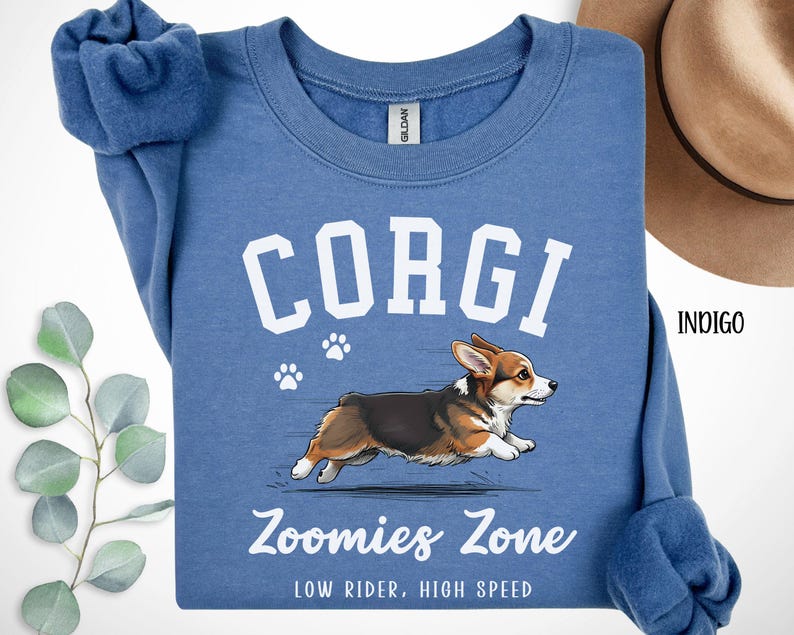 May include: Indigo blue sweatshirt with the word "CORGI" in white block letters above a running corgi illustration. The text "Zoomies Zone" and "LOW RIDER, HIGH SPEED" are printed below the dog. Paw prints are also included.