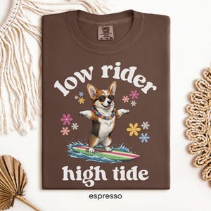 May include: Espresso-colored t-shirt with a graphic of a corgi wearing sunglasses and a lei, surfing on a surfboard. The text "low rider high tide" is above and below the corgi illustration. The shirt has a crew neck.