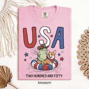 Puede incluir: Camiseta rosa claro con un dise&ntilde;o gr&aacute;fico que dice "USA" en rojo, blanco y azul. Una rana con sombrero de vaquero y un corgi con pa&ntilde;uelo est&aacute;n en una boya. El texto "TWO HUNDRED AND FIFTY" y "blossom" est&aacute;n en la parte inferior.