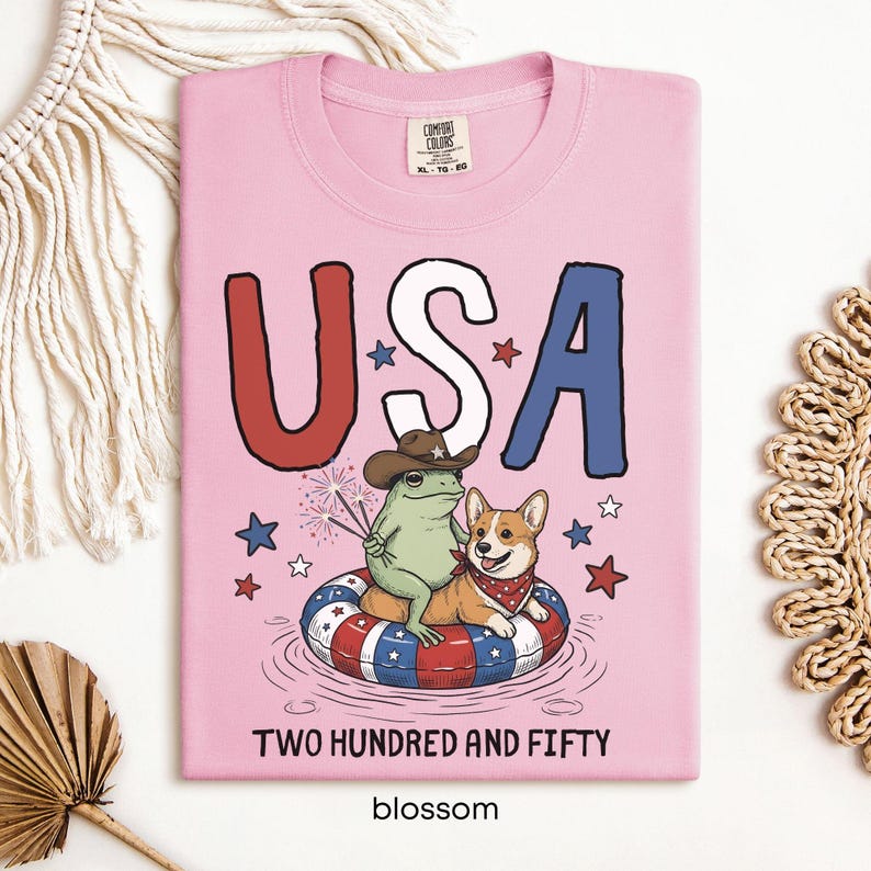 Puede incluir: Camiseta rosa claro con un dise&ntilde;o gr&aacute;fico que dice "USA" en rojo, blanco y azul. Una rana con sombrero de vaquero y un corgi con pa&ntilde;uelo est&aacute;n en una boya. El texto "TWO HUNDRED AND FIFTY" y "blossom" est&aacute;n en la parte inferior.