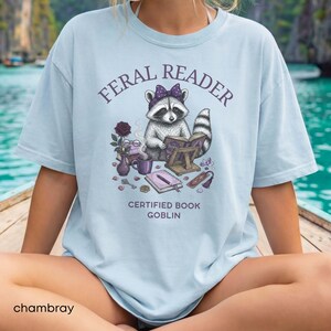May include: Light blue chambray t-shirt with the text "FERAL READER" above an illustration of a raccoon reading a book. The raccoon wears a purple bow and is surrounded by books, a rose, and a mug. The text "CERTIFIED BOOK GOBLIN" is below the illustration.