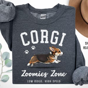 May include: Dark heather gray sweatshirt with the word "CORGI" in white block letters above a graphic of a running corgi dog. The text "Zoomies Zone" and "LOW RIDER, HIGH SPEED" are below the dog illustration. The sweatshirt has a crew neck and long sleeves.