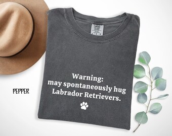 Puede incluir: Camiseta gris oscuro con el texto blanco "Warning: may spontaneously hug Labrador Retrievers" y una huella de pata. Un sombrero beige está en la esquina superior izquierda. Una ramita de hojas verdes está a la derecha.