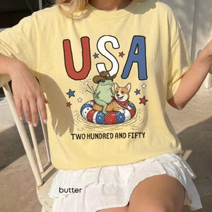 Puede incluir: Una camiseta amarilla clara con la palabra "USA" en rojo y azul, con una rana y un perro de dibujos animados en una c&aacute;mara de aire. La camiseta tambi&eacute;n tiene el texto "TWO HUNDRED AND FIFTY" y la palabra "butter" en la parte inferior.