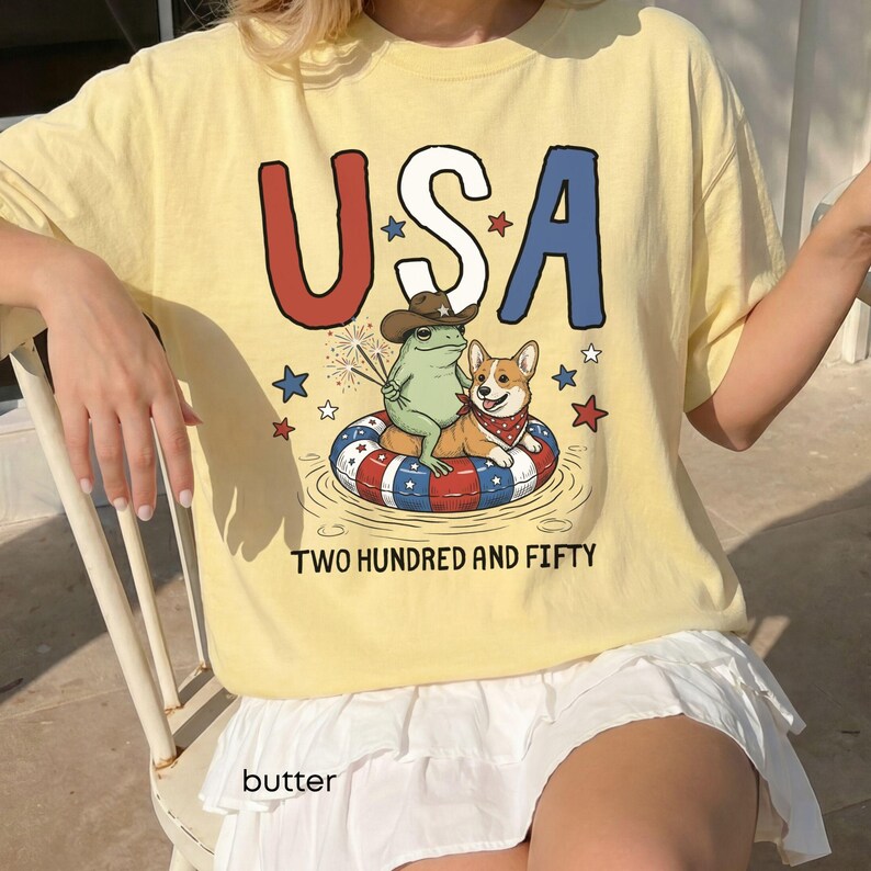 Puede incluir: Una camiseta amarilla clara con la palabra "USA" en rojo y azul, con una rana y un perro de dibujos animados en una c&aacute;mara de aire. La camiseta tambi&eacute;n tiene el texto "TWO HUNDRED AND FIFTY" y la palabra "butter" en la parte inferior.