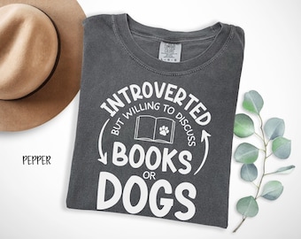 Può includere: Una maglietta grigio scuro con la scritta bianca "INTROVERTED BUT WILLING TO DISCUSS BOOKS OR DOGS". Il design include un libro e un'impronta di zampa. Un cappello beige e un rametto di verde sono sullo sfondo.