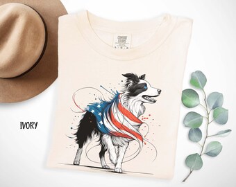 May include: A white t-shirt with a black and white border collie dog wearing an American flag. The dog is standing on a cream colored background with red, white, and blue paint splatters.