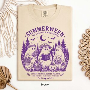 May include: A beige t-shirt featuring a purple graphic of three ghosts in a forest. The text reads "SUMMERWEEN," "WILD HEARTS & WILDFLOWERS," "GHOST STORIES," and "COTTAGE NIGHTS & CURSED DELIGHTS - PICK FLOWERS - READ BOOKS - FIND MAGIC."