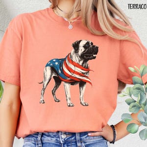 May include: A terracotta colored t-shirt with a graphic of a dog wearing an American flag. The dog is a Bullmastiff breed.