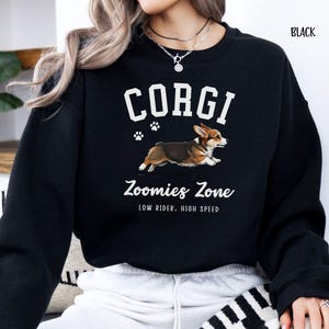 May include: Black sweatshirt featuring a graphic of a running corgi dog with the word "CORGI" above it. Below the dog is the text "Zoomies Zone" and "LOW RIDER, HIGH SPEED". The word "BLACK" is in the upper right corner.