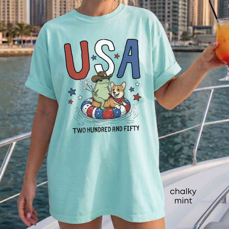 Puede incluir: Una camiseta verde menta con la palabra "USA" en rojo, blanco y azul. La camiseta presenta un gr&aacute;fico de una rana y un corgi en un flotador patri&oacute;tico, con el texto "TWO HUNDRED AND FIFTY".