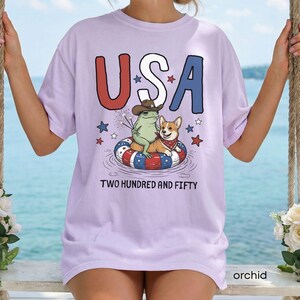 Puede incluir: Camiseta color orqu&iacute;dea con la palabra "USA" en rojo y azul, con una rana con sombrero de vaquero y un corgi en un flotador patri&oacute;tico. El texto "TWO HUNDRED AND FIFTY" est&aacute; debajo.