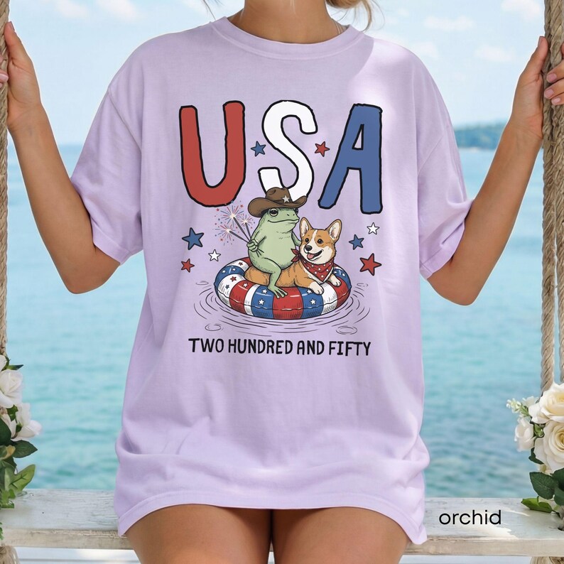 Puede incluir: Camiseta color orqu&iacute;dea con la palabra "USA" en rojo y azul, con una rana con sombrero de vaquero y un corgi en un flotador patri&oacute;tico. El texto "TWO HUNDRED AND FIFTY" est&aacute; debajo.