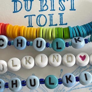 Op de afbeelding: Een verzameling kleurrijke kralenarmbanden. Eén armband heeft regenboogkleurige schijfkralen. Andere armbanden hebben kralen met letters die "SCHULKIND" en "HULKIND" spellen, met hartvormige kralen. De achtergrond heeft de tekst "DU BIST TOLL".