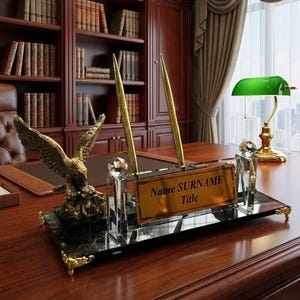 May include: A desk set featuring a gold eagle statue, a pen holder with two gold pens, and a nameplate. The nameplate reads "Name SURNAME Title". The set is on a mirrored base with gold accents. A green desk lamp is visible.