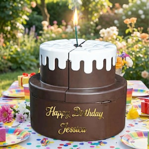 Könnte beinhalten: Zweistöckige, braune Geburtstagstorte mit weißer Glasur und einer brennenden Kerze. Auf der Torte steht in goldener Schrift "Happy Birthday Jessica". Die Torte steht auf einem Tisch mit bunten Partyutensilien und Blumen im Hintergrund.