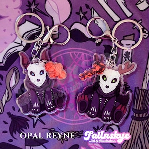 May include: Two keychains with silver clasps. Each features a purple and black creature with a skull-like face and a red accent. The text "OPAL REYNE" and "Fallnskeye Art & Illustration" are visible.