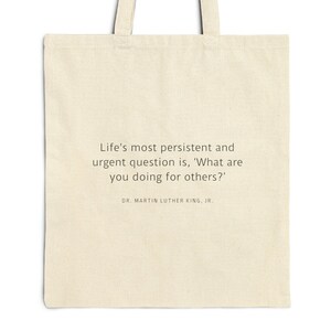 Puede incluir: Una bolsa de tela beige con una cita de Martin Luther King Jr. que dice: "La pregunta más persistente y urgente es: '¿Qué estás haciendo por los demás?'"