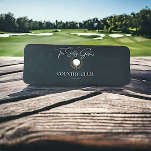 May include: A black card with white text that reads "The Shitty Golfers Country Club" with a gold golf ball and laurel wreath logo.