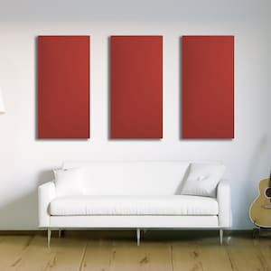 May include: Three rectangular, solid red wall panels are mounted on a white wall above a white sofa with two pillows. A floor lamp and a guitar are also in the room. The floor is made of wood.