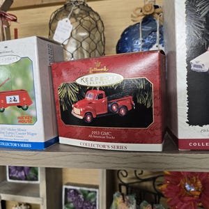 May include: A red Hallmark Keepsake ornament box featuring a 1953 GMC All-American Trucks miniature. The box is red with gold lettering and a picture of the truck. The box is part of the Collector's Series.