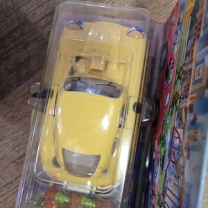 Puede incluir: Un coche de juguete amarillo en un embalaje de plástico transparente. El coche tiene una forma redondeada con detalles azules en los faros. El embalaje incluye gráficos coloridos de una escena urbana.