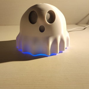 May include: A white ghost-shaped speaker with a blue light emanating from the bottom. The speaker has two black speaker grilles for the eyes and a round black speaker grille for the mouth.