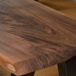 May include: Close-up of a wooden bench with a natural edge. The bench features a rich, dark brown wood grain and a smooth, polished surface. The legs are black metal. The bench is a piece of furniture.