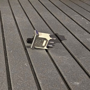 以下が含まれることがあります： 銀色の金属製の接点と青いボタンが付いた、小さく白い長方形のトグルスイッチ。スイッチは黒と黄色のテクスチャのある表面にあります。