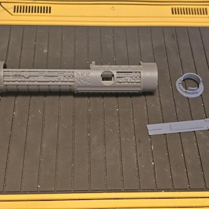 Op de afbeelding: Een grijze, 3D-geprinte lichtzwaardgreep met ingewikkelde details, samen met verschillende losse componenten. De greep heeft een getextureerde grip en verschillende geometrische patronen. De onderdelen zijn gerangschikt op een zwart oppervlak met gele accenten.