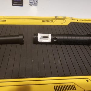 Op de afbeelding: Een gedemonteerde zwarte lichtzwaardgreep met een wit intern onderdeel en een aparte zwarte handgreep. Er is ook een witte rubberen ring aanwezig. Het lichtzwaard ligt op een zwart oppervlak met gele accenten.