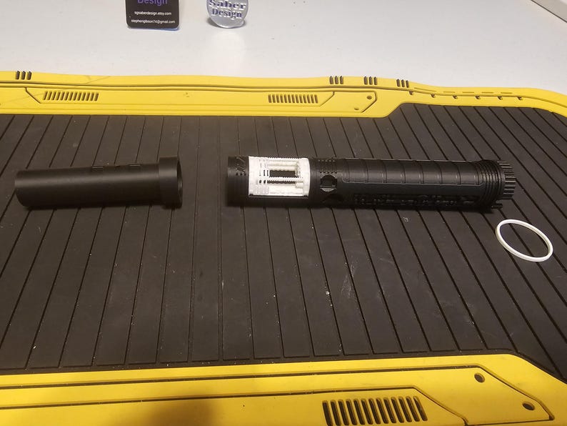 Op de afbeelding: Een gedemonteerde zwarte lichtzwaardgreep met een wit intern onderdeel en een aparte zwarte handgreep. Er is ook een witte rubberen ring aanwezig. Het lichtzwaard ligt op een zwart oppervlak met gele accenten.