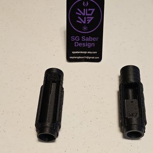 May include: Two black, cylindrical components with detailed ridges and grooves, possibly parts of a larger assembly. A business card with the text "SG Saber Design" and contact information is also present.