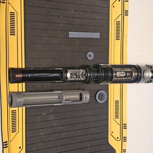 Op de afbeelding: Onderdelen van een gedemonteerde lichtzwaard, waaronder een zwarte en zilveren handgreep, een grijs cilindrisch gedeelte en diverse elektronische onderdelen. De onderdelen liggen op een zwarte ondergrond met gele randdetails.