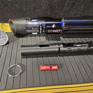 Könnte beinhalten: Ein schwarzer Lichtschwertgriff mit einer silbernen Klinge, eine schwarze Lichtschwertverlängerung, ein rotes Plastikteil und ein silberner Ring. Der Lichtschwertgriff hat einen blauen Knopf und einen schwarzen Aufkleber mit dem Text "SG Saber Design".