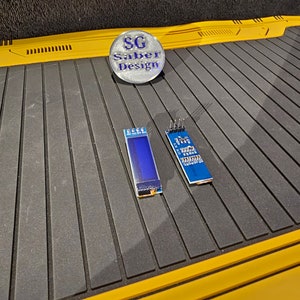 May include: Two blue circuit boards with small blue screens and silver connectors. The boards are on a black and yellow surface. A silver button with the text "SG Saber Design" is in the upper left corner of the image.