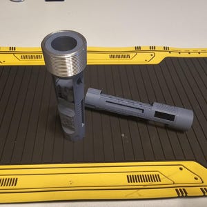 Puede incluir: Dos objetos cilíndricos grises con diseños detallados. Uno tiene una parte superior metálica roscada. El otro es un cilindro más pequeño y alargado. Los objetos están sobre una superficie negra y amarilla.