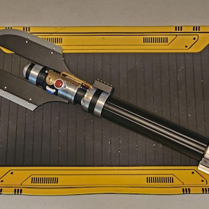 Op de afbeelding: Een zwart en zilverkleurig lichtzwaard met een uniek bladontwerp. Het handvat heeft zilveren en zwarte accenten, een rode knop en een goudkleurig detail. Het blad is donkergrijs van kleur en heeft een opvallende vorm.