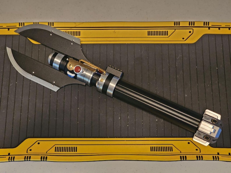 Op de afbeelding: Een zwart en zilverkleurig lichtzwaard met een uniek bladontwerp. Het handvat heeft zilveren en zwarte accenten, een rode knop en een goudkleurig detail. Het blad is donkergrijs van kleur en heeft een opvallende vorm.