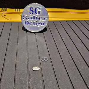 May include: Two small electronic switches, one black and one white, on a textured gray surface. The switches are rectangular and have a small button on the top. The surface is made of a material that looks like rubber or plastic. The background is a yellow surface with a circular sticker that says "SG Saber Design".